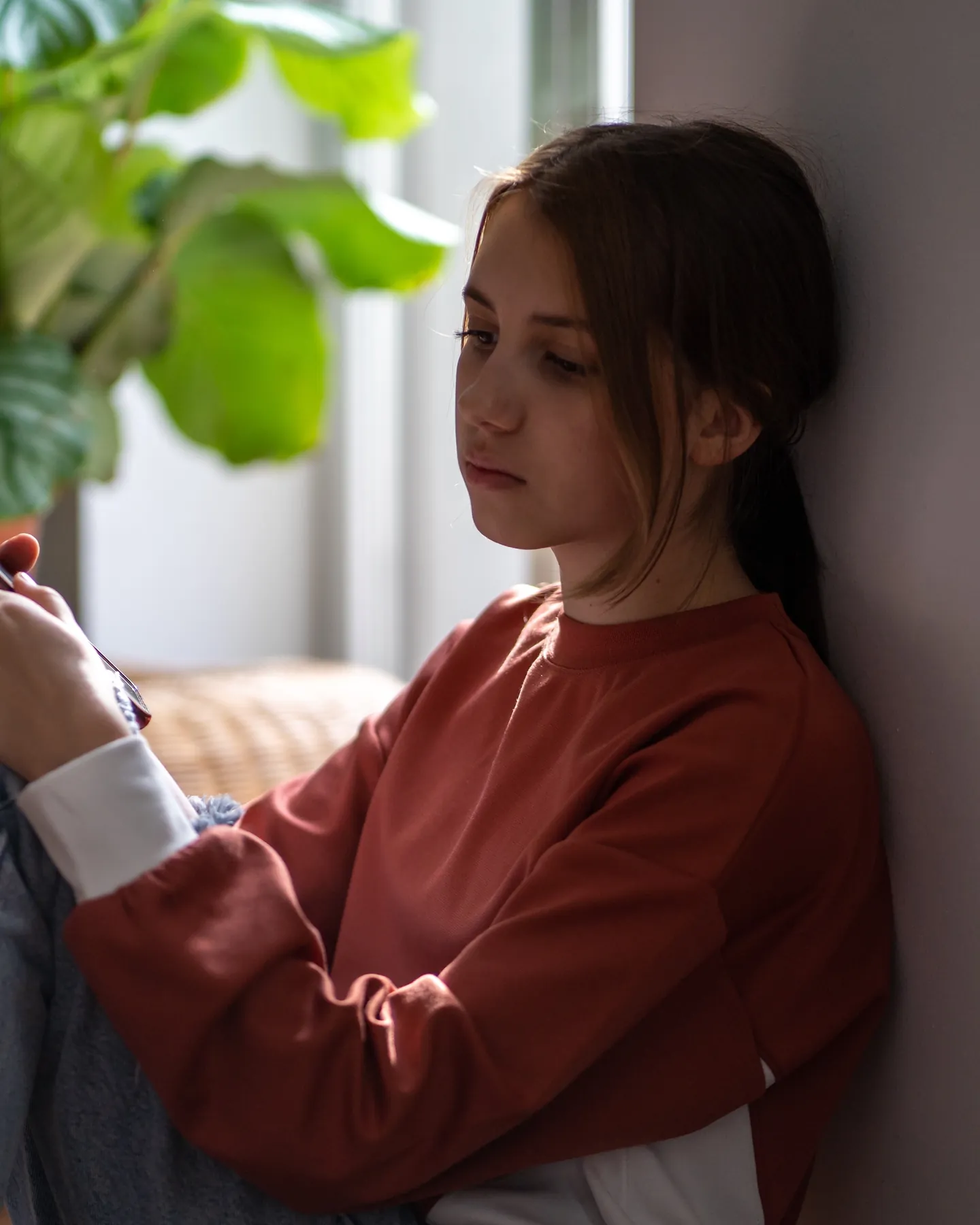 A teenage girl in a rust sweatshirt sits against a wall by a window, looking down at her phone.