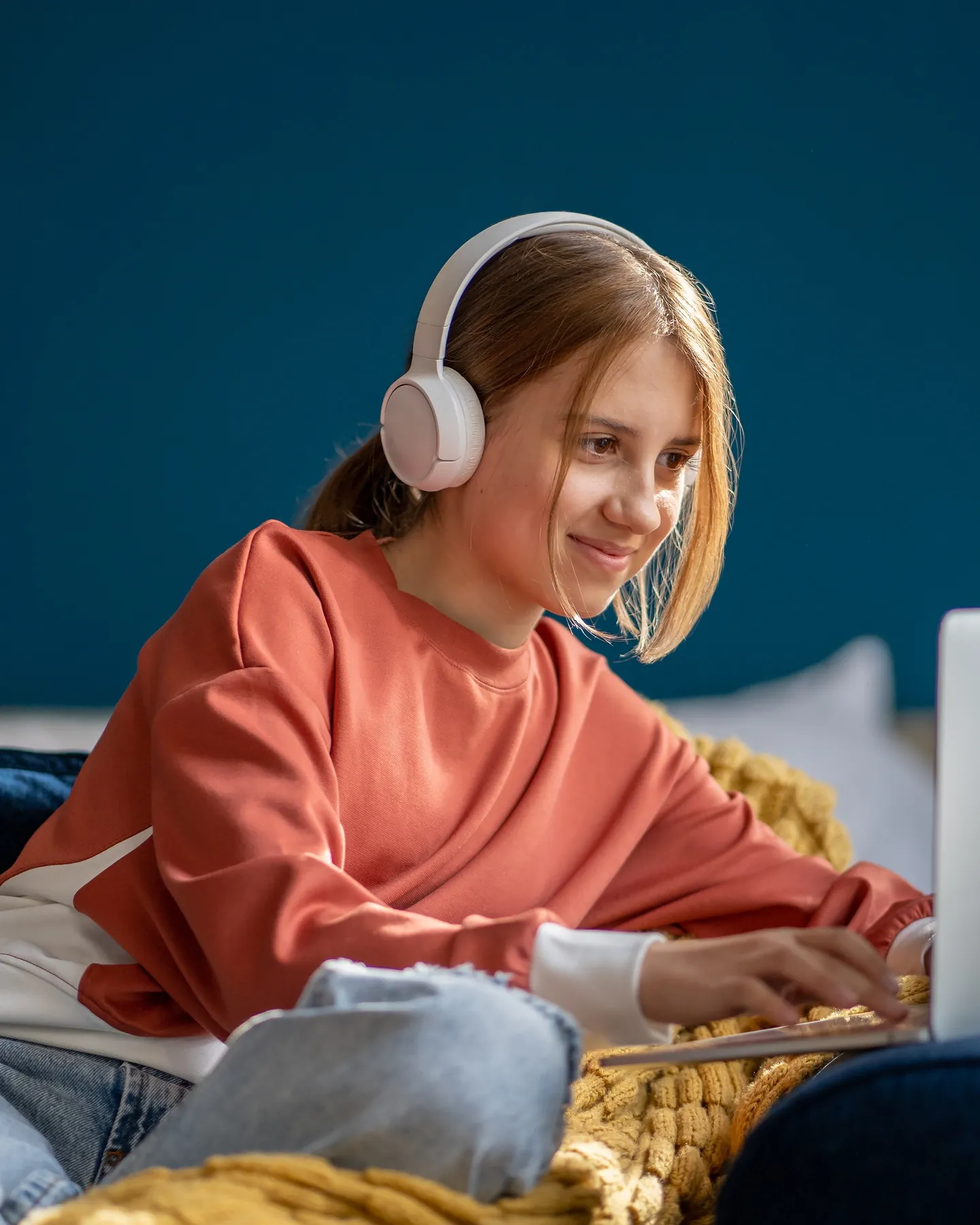 A teenage girl wearing headphones smiles at a laptop screen while lying on a bed with a yellow blanket.